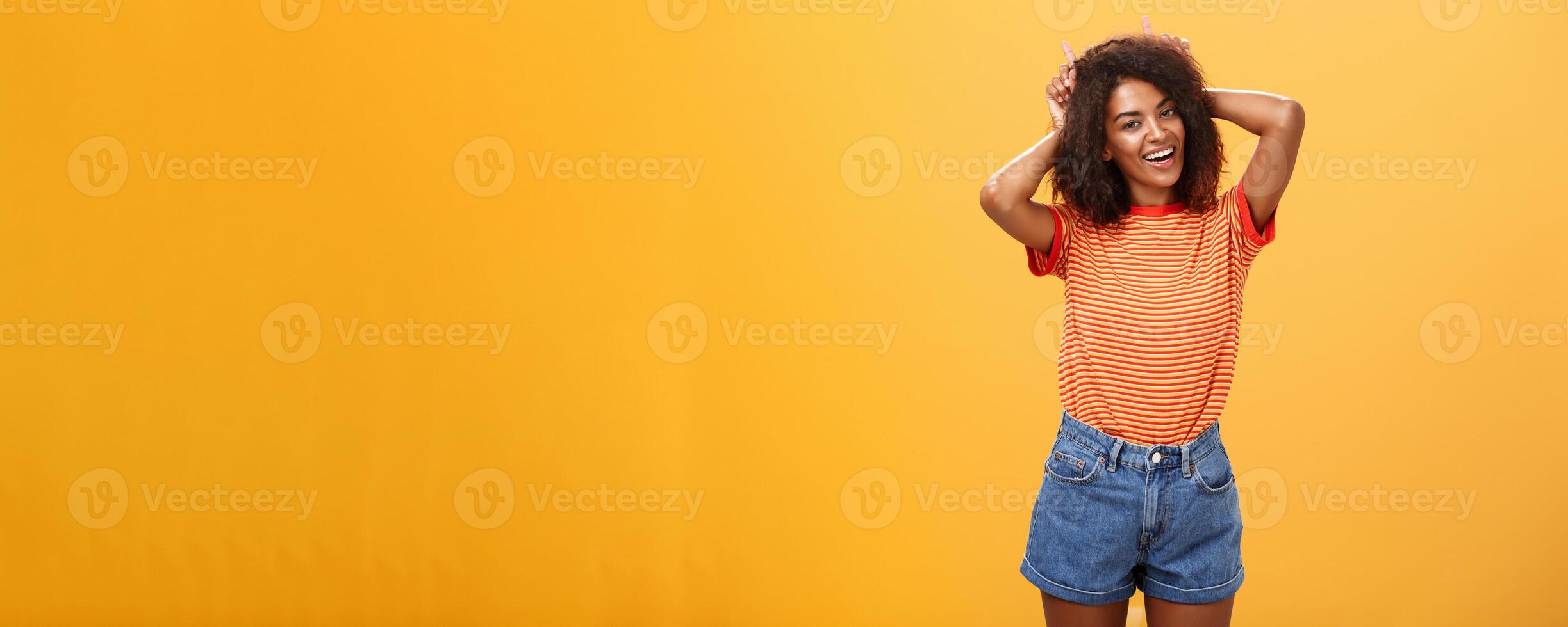 Rebellious playful and joyful african american girl with curly hairstyle making devil horns on head with index fingers smiling broadly showing daredevil character and charisma over orange wall photo