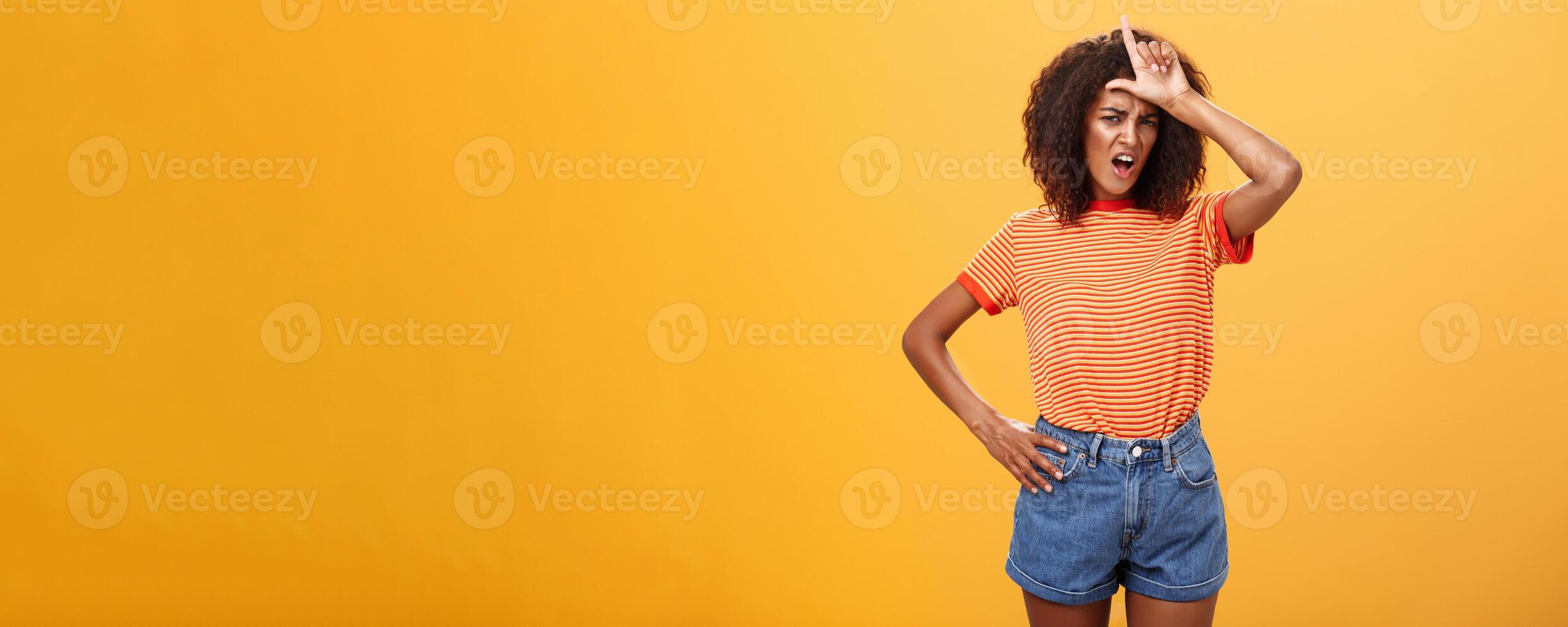 Arrogant and confident woman not talk with losers. Mocking self-assured stylish african american female with curly hair in trendy clothes frowning scolding nerd, showing letter l on forehead photo
