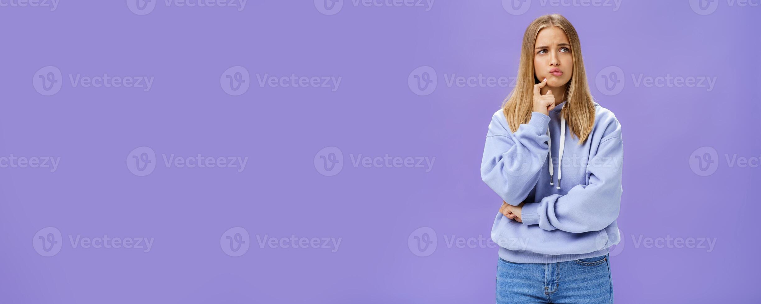 Silly concerned girlfriend trying think up how solve problem standing thoughtful with troubled expression touching jaw pouting, frowning staring right, thinking making up decision over purple wall photo