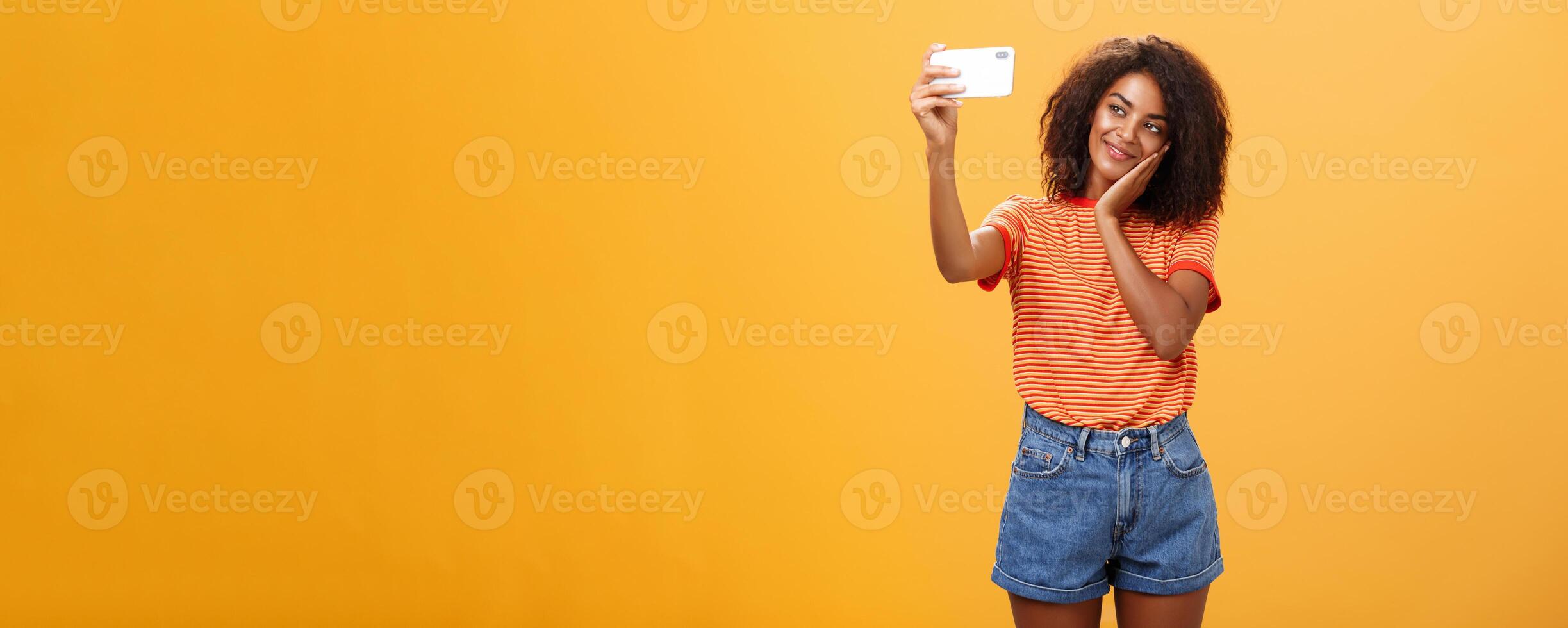 Woman made make-up all day to post new photo online taking selfie on brand new smartphone making cute face leaning head on palm smiling gently at device screen posing over orange background