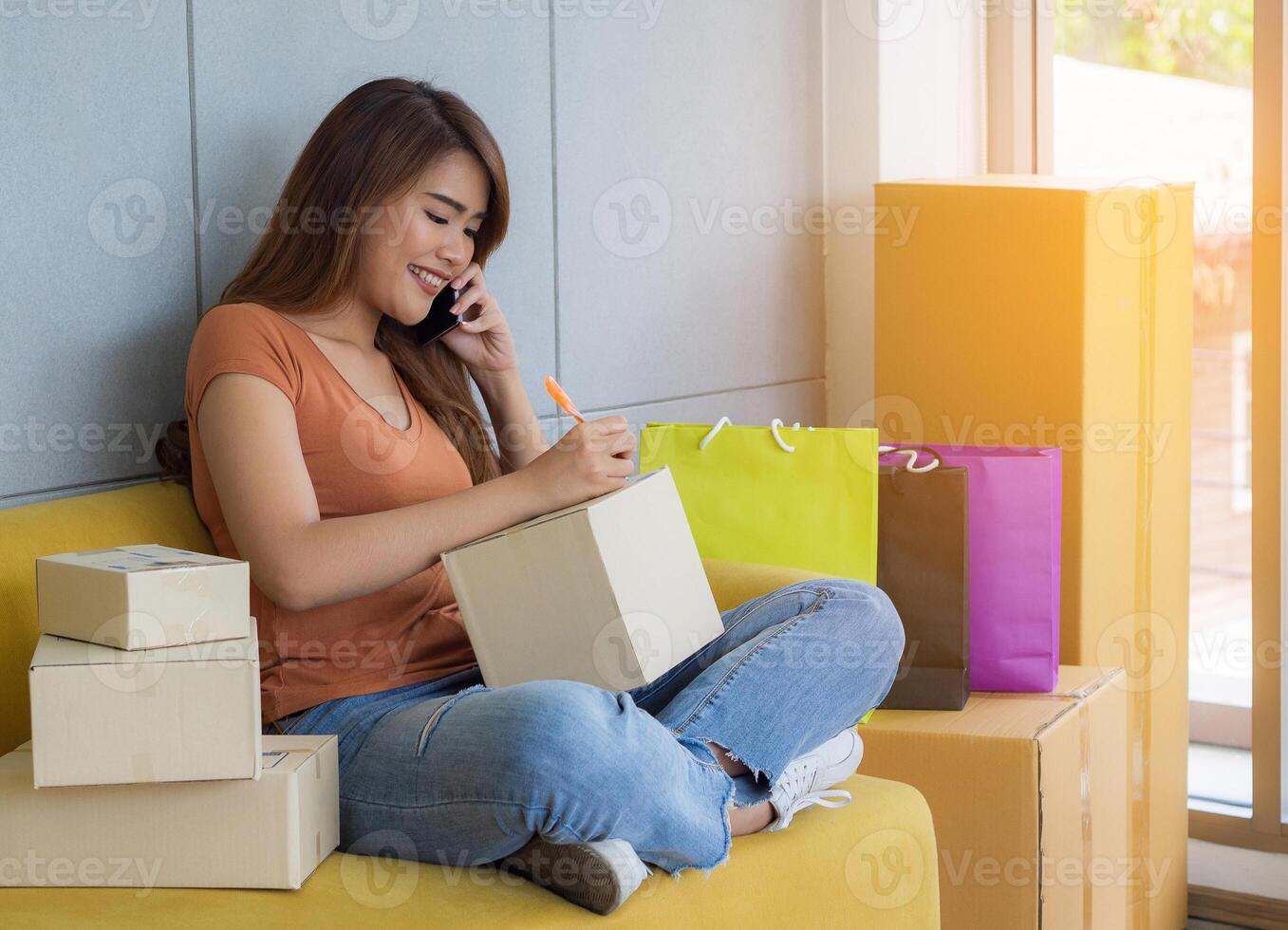 asiático mujer de negocios es un comercio electrónico empresario utilizando un teléfono inteligente y escribe cliente habla a en un paquete o empaquetar caja mientras sentado en un sofá en el oficina. concepto de comercio electrónico foto
