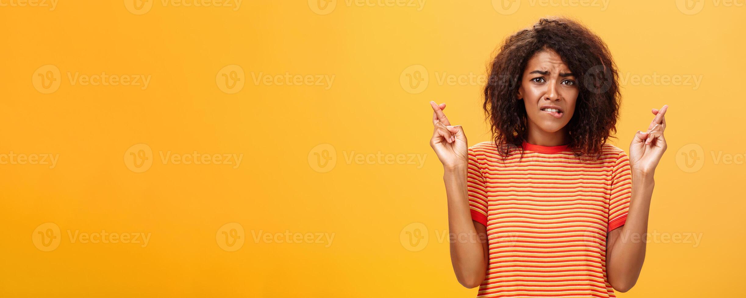 Waist-up shot of concerned troubled nervous african american woman with curly hairstyle biting lower lip anxiously frowning crossing fingers for good luck hopefully praying for dream come true photo