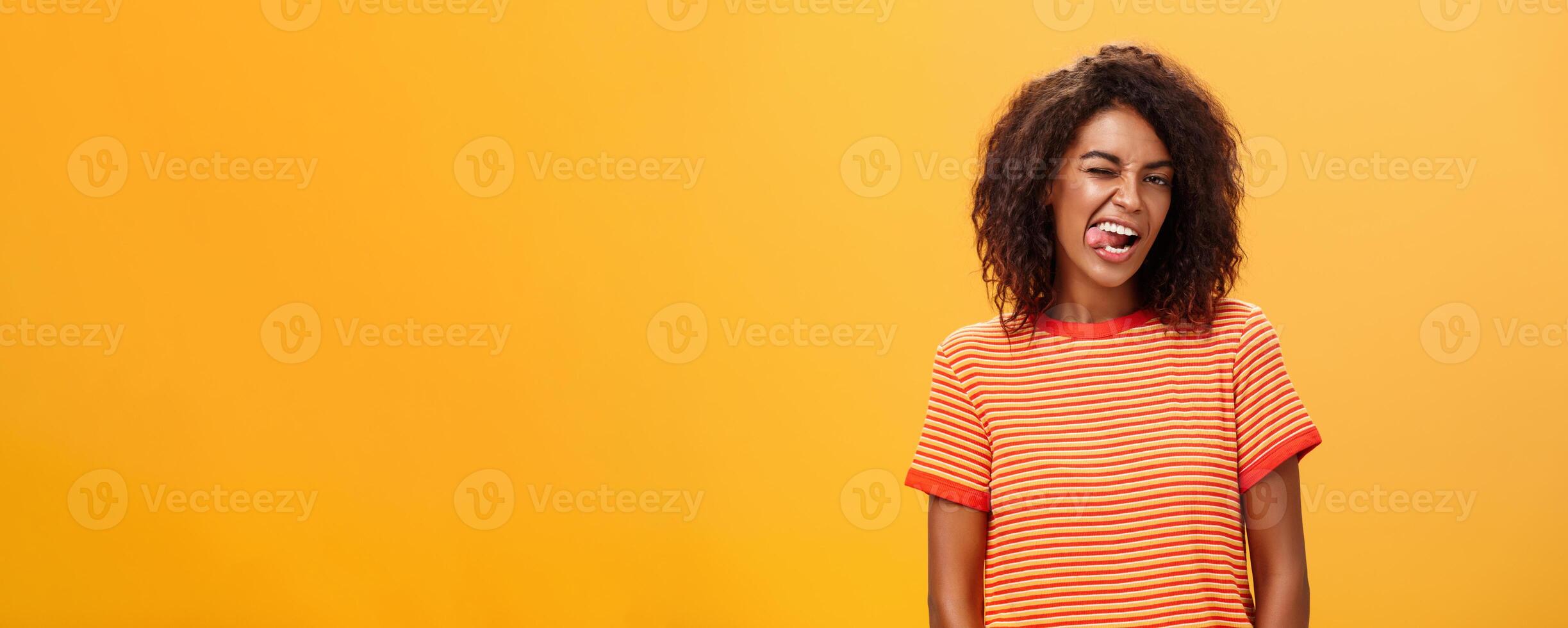Portrait of daring and emotive confident flirty woman with afro hairstyle winking joyfully showing tongue posing carefree and enthusiastic against orange background flirting with hot guy photo