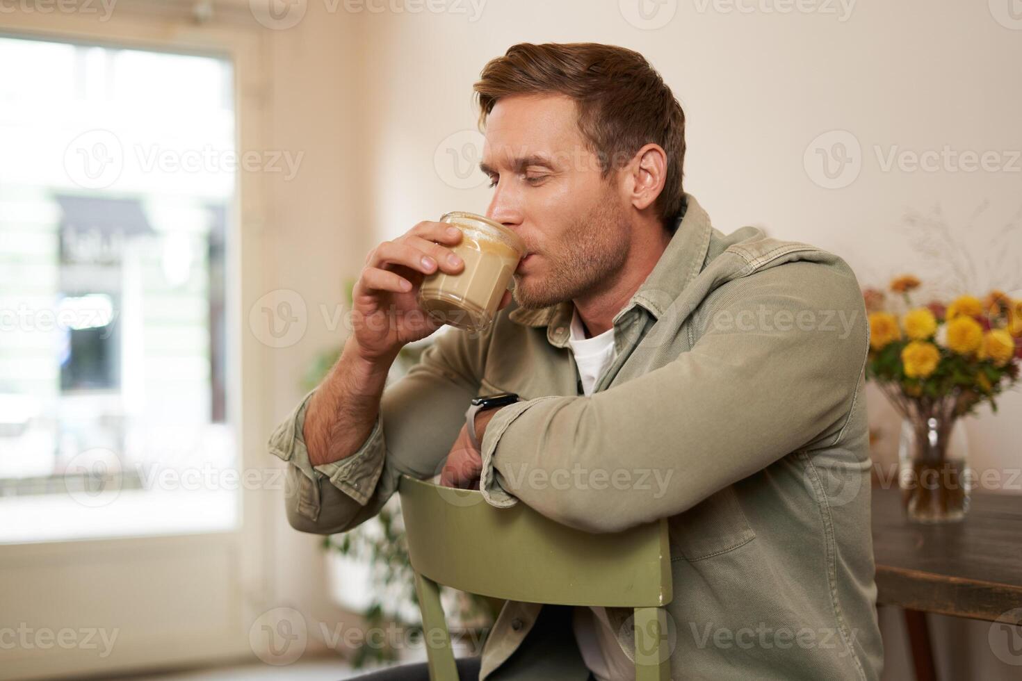 Portrait of handsome blond man in cafe, sits on chair, enjoys the taste of delicious freshly brewed cup of coffee photo