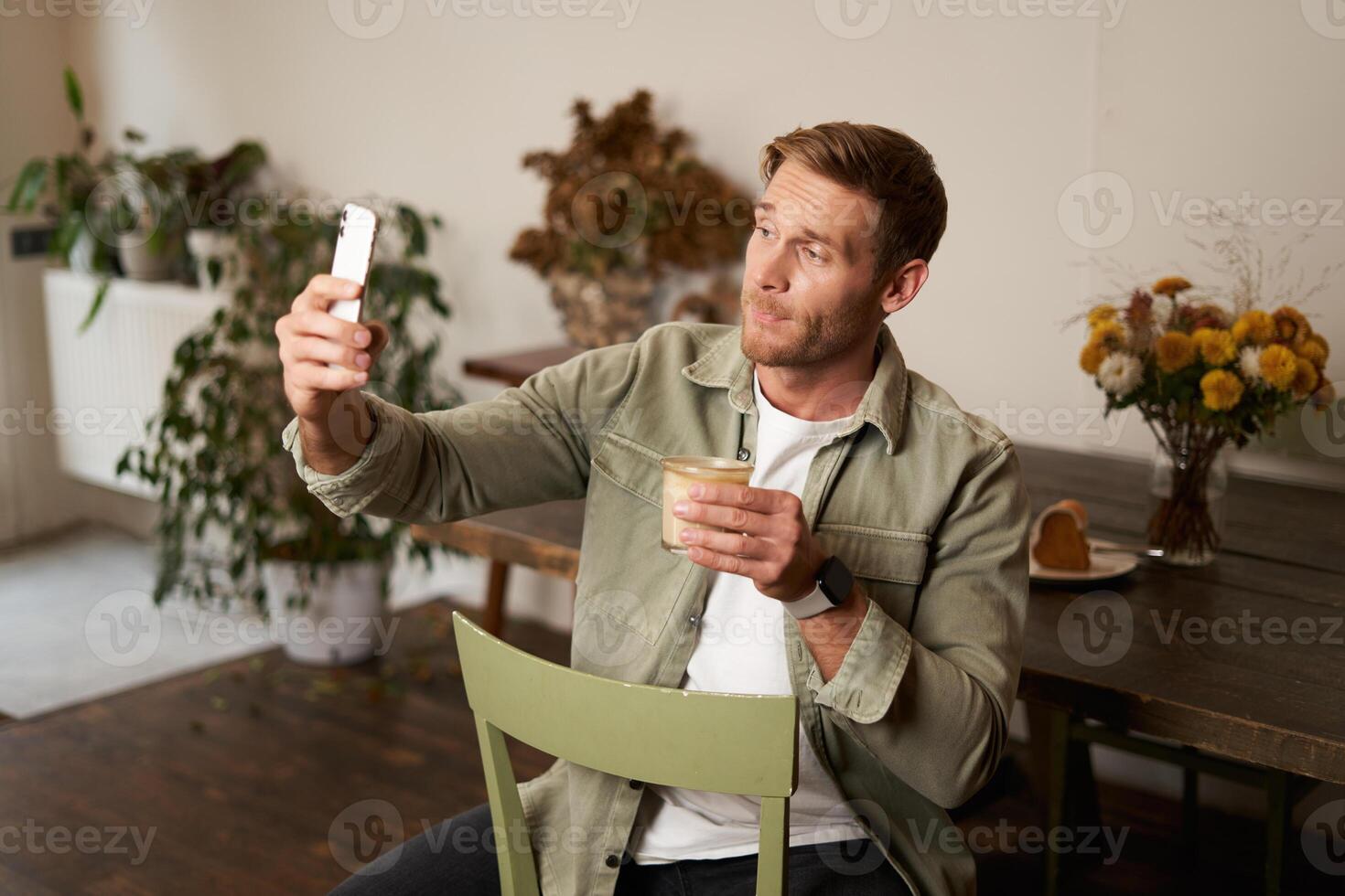 Portrait of young handsome man takes slefie on his mobile phone app, sits in cafe with glass of coffee, posting on social media application photo