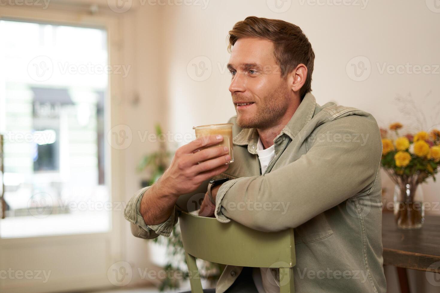 Close up portrait of handsome man, sitting in cafe on chair, holding glass of coffee, drinking cappuccino and smiling. Concept of lifestyle and leisure photo