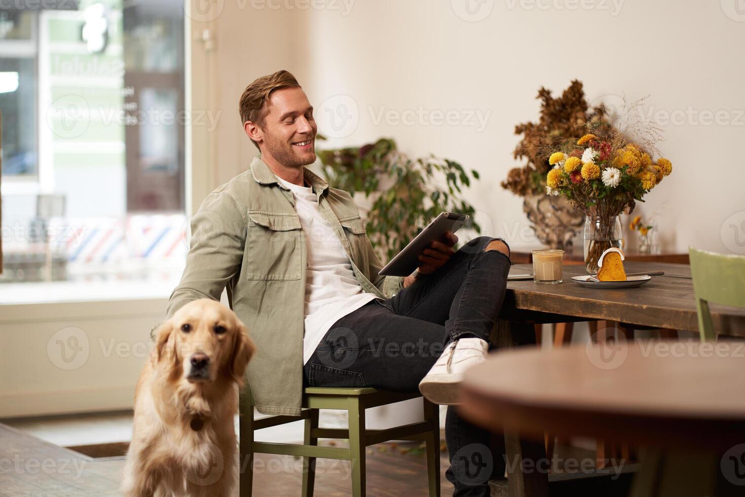 Portrait of handsome young man sits in cafe with his dog, petting golden retriever, reading news on tablet, relaxing with cup of coffee photo