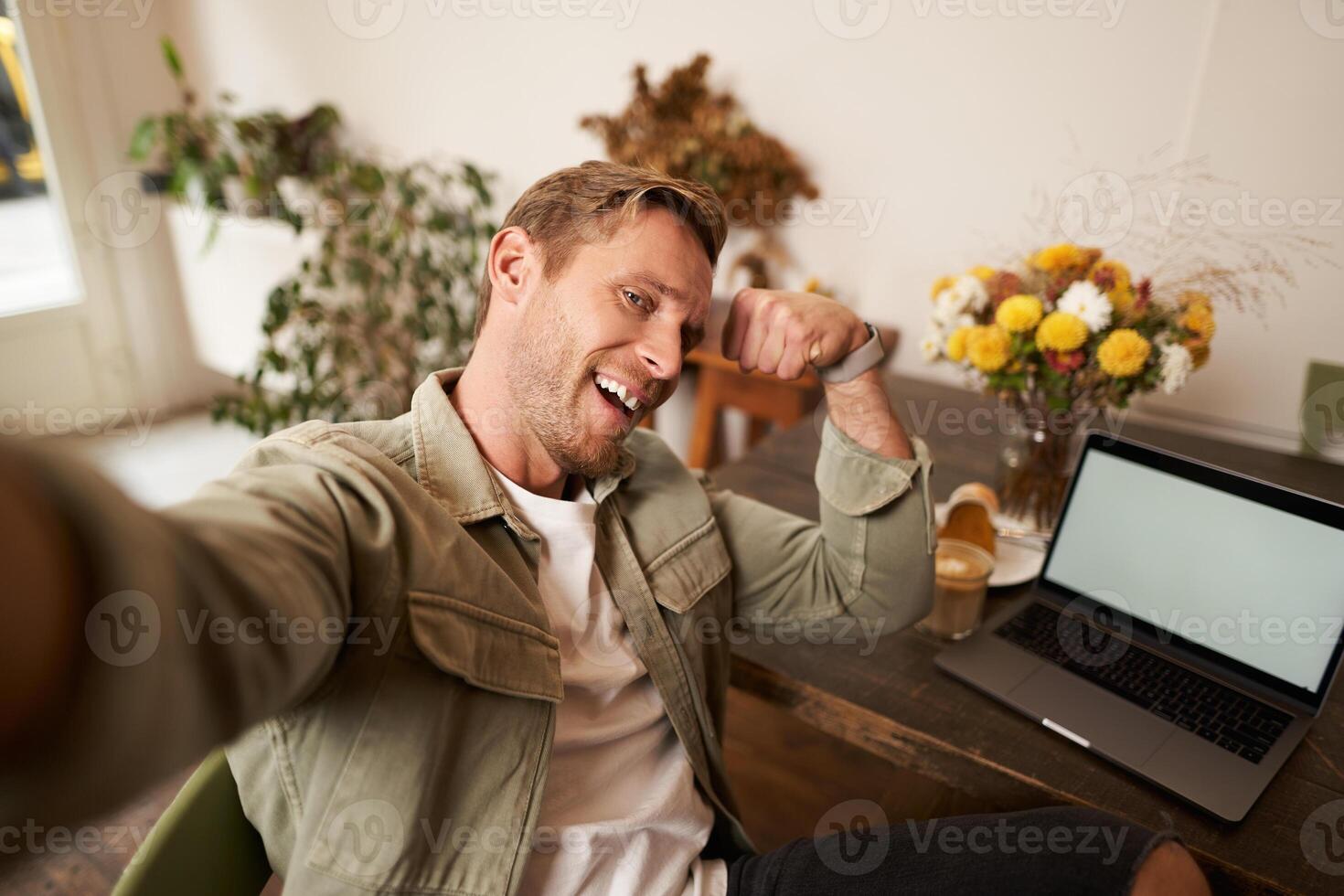 Portrait of handsome and confident man taking a selfie, flexing biceps, shows-off his muscles, sits in cafe in front of laptop, smiling at camera photo