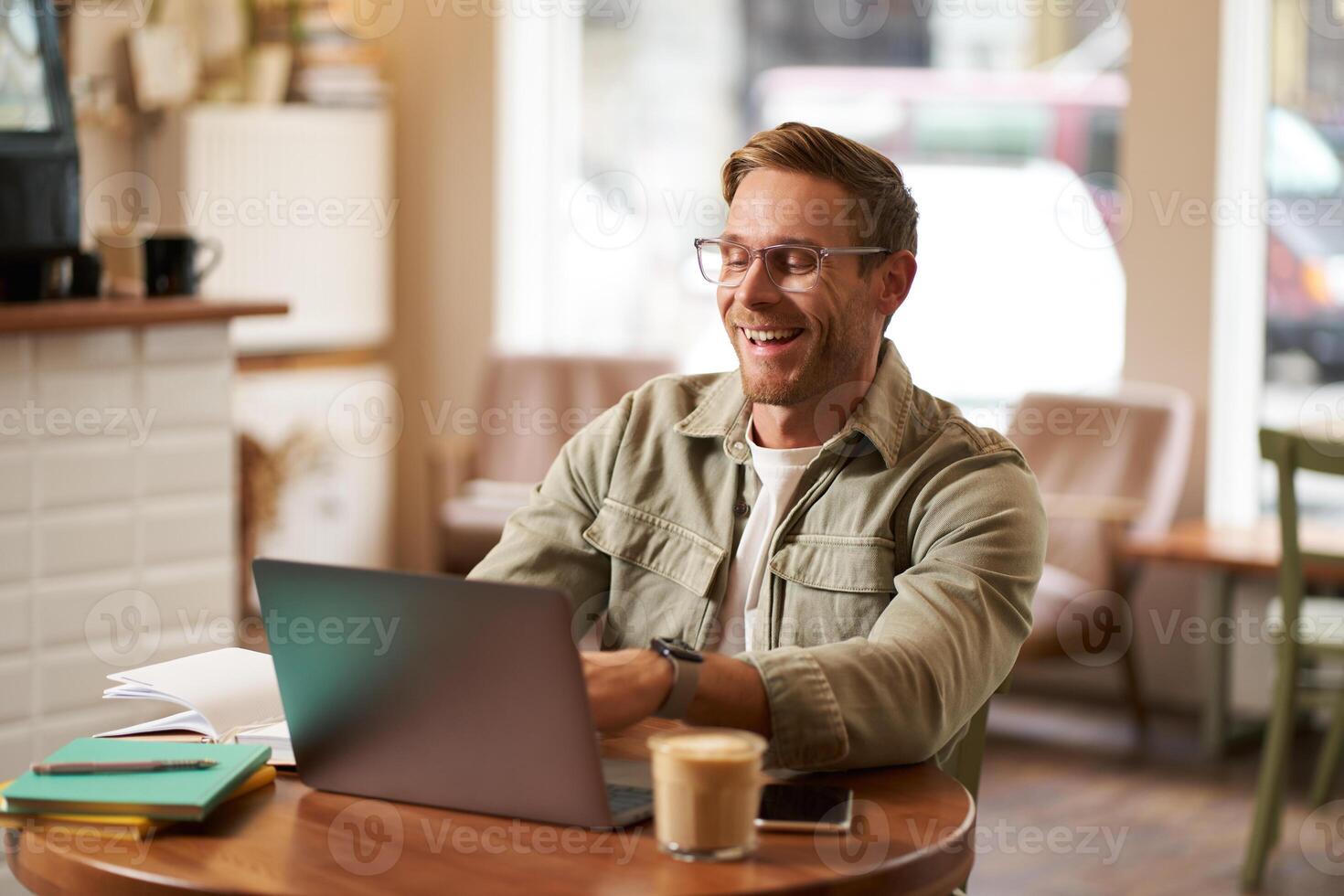 Image of young digital nomad, man in glasses sits in cafe, works from coffee shop, uses laptop in co-working space, wears glasses, drinks his beverage photo