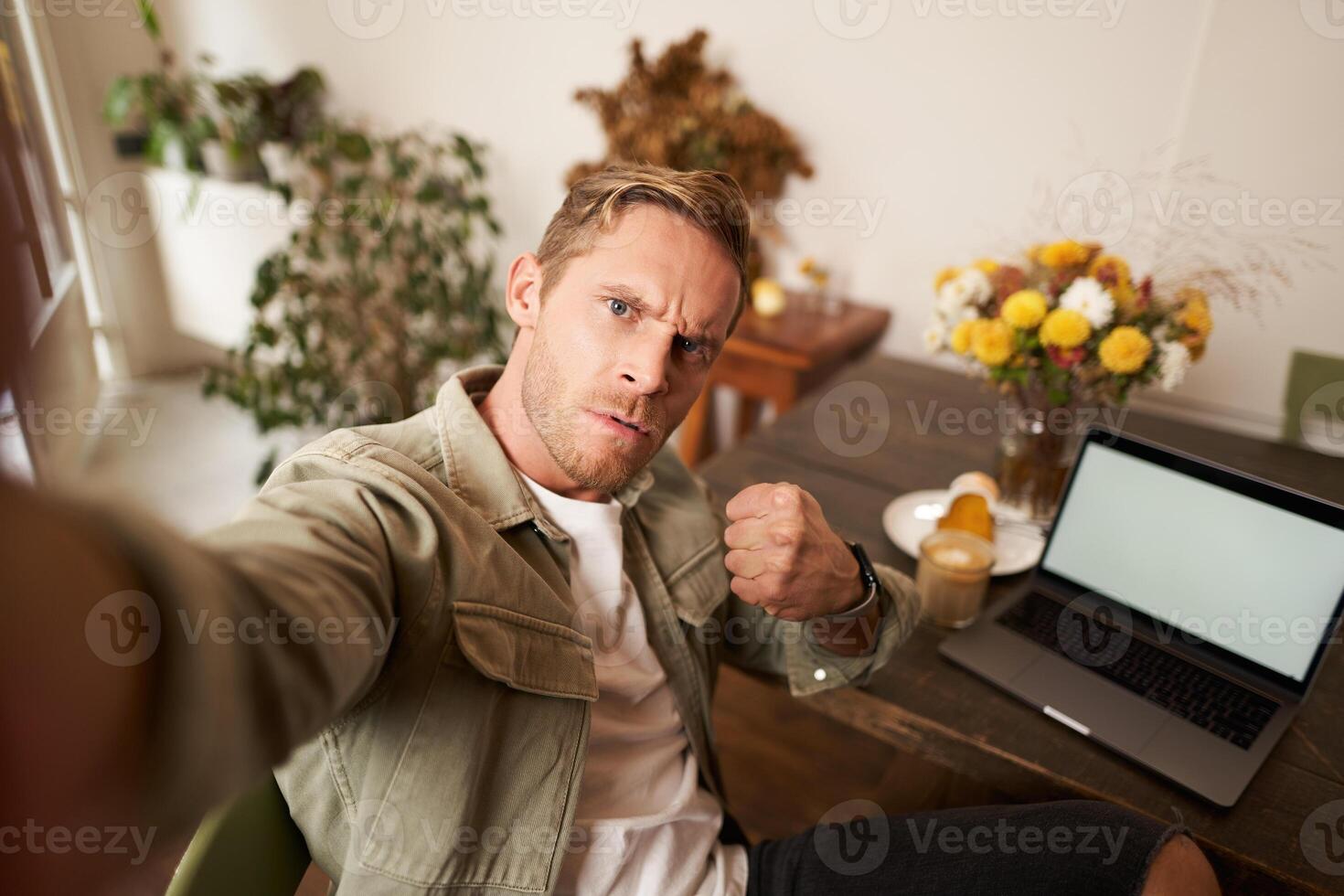 retrato de grave hombre toma selfie con puño y amenazante rostro, se sienta en cafetería, poses en café tienda en frente de mesa con ordenador portátil y vaso de café foto