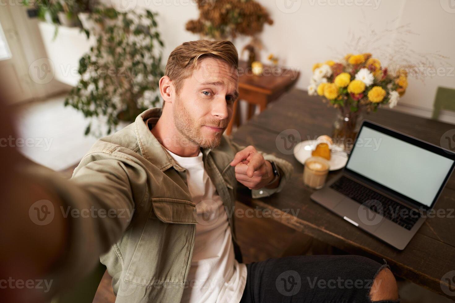 Handsome and confident young man taking selfie on mobile phone, pointing finger at camera, sitting in cafe with laptop, working from coffee shop, looking sassy photo