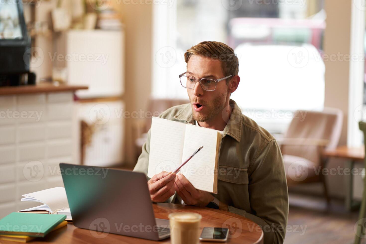 Portrait of handsome young man giving lessons online, showing exercises in his notebook, looking at laptop, recording , sitting in cafe, quite co-working space photo