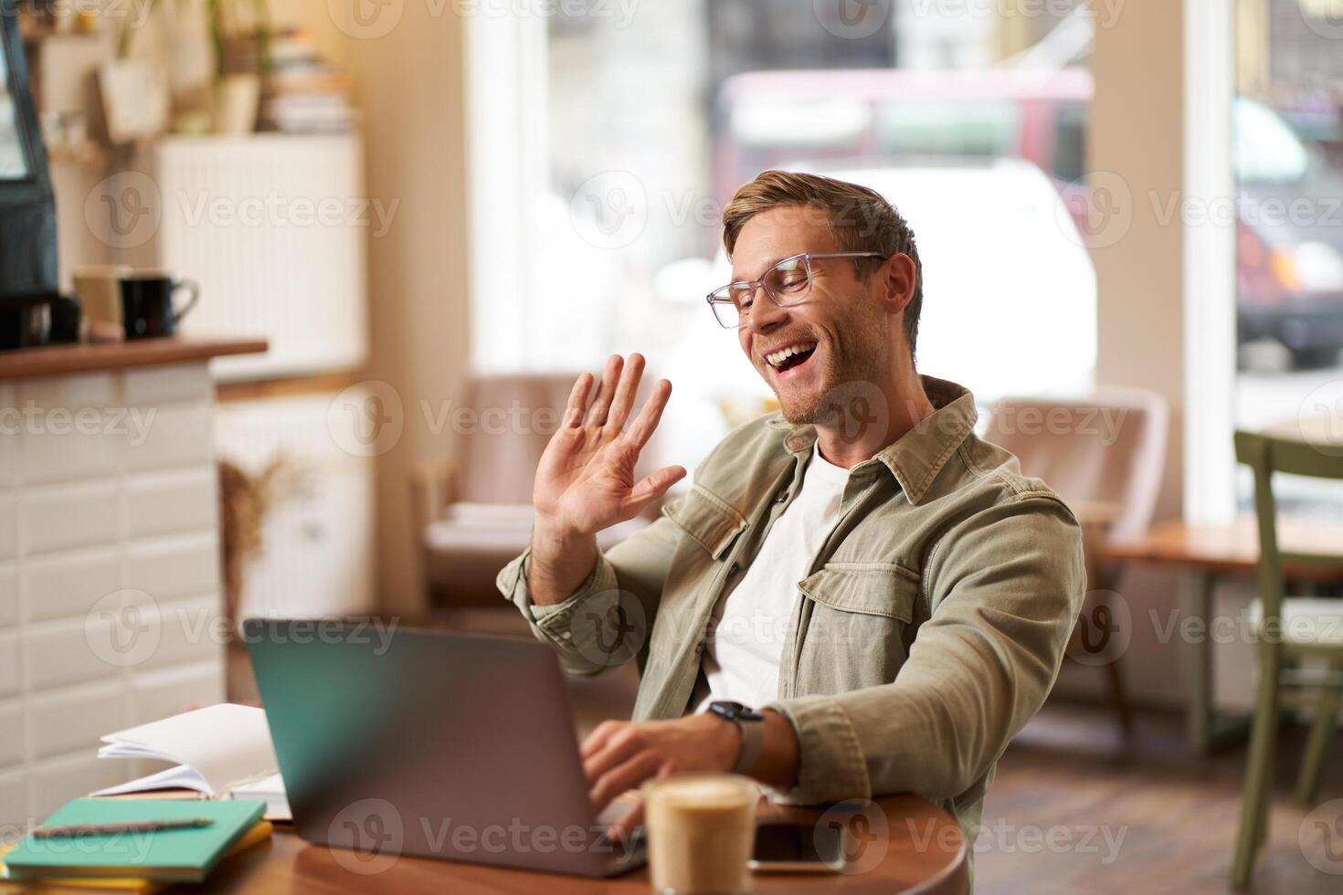 Portrait of handsome, smiling young man, online tutor, businessman working in cafe remotely, waving hand at laptop, connects to chat or meeting photo