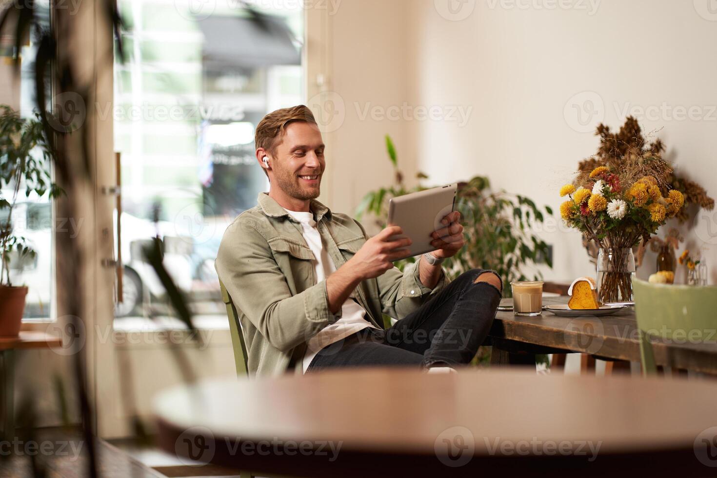 Portrait of handsome happy guy, young man sits in cafe, watching s on digital tablet, wearing wireless headphones, laughing and smiling, spending time in coffee shop photo