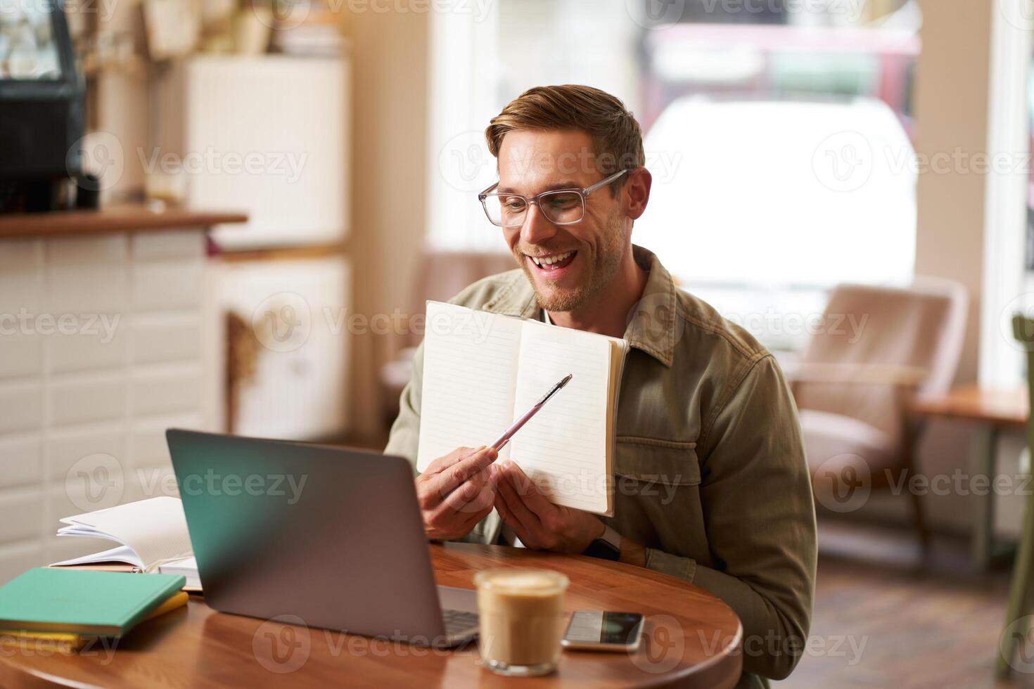 Portrait of young handsome man in glasses, private tutor teaching student online, pointing at his notebook, showing something, chats via laptop application, working remotely in cafe photo