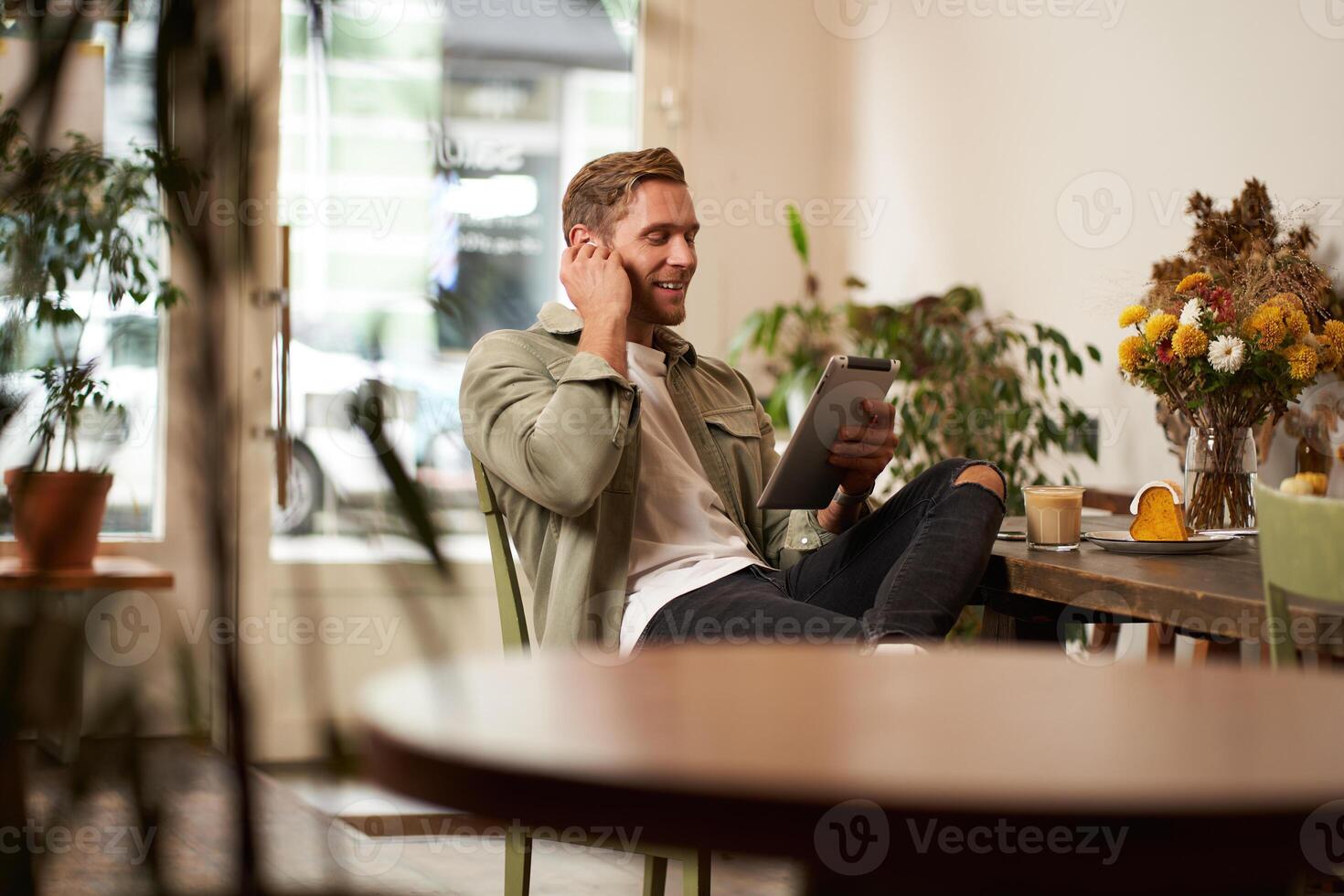 Portrait of handsome happy guy, young man sits in cafe, watching s on digital tablet, wearing wireless headphones, laughing and smiling, spending time in coffee shop photo