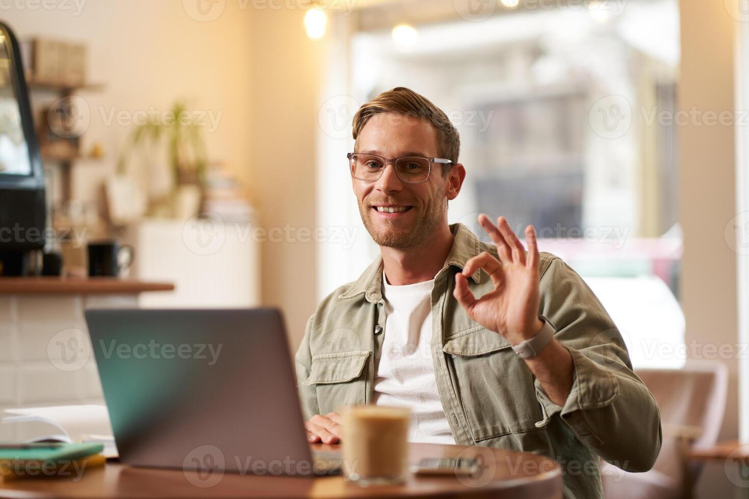 Portrait of handsome businessman in glasses, showing okay hand sign, sitting with laptop in cafe, working remotely, recommends co-working space photo