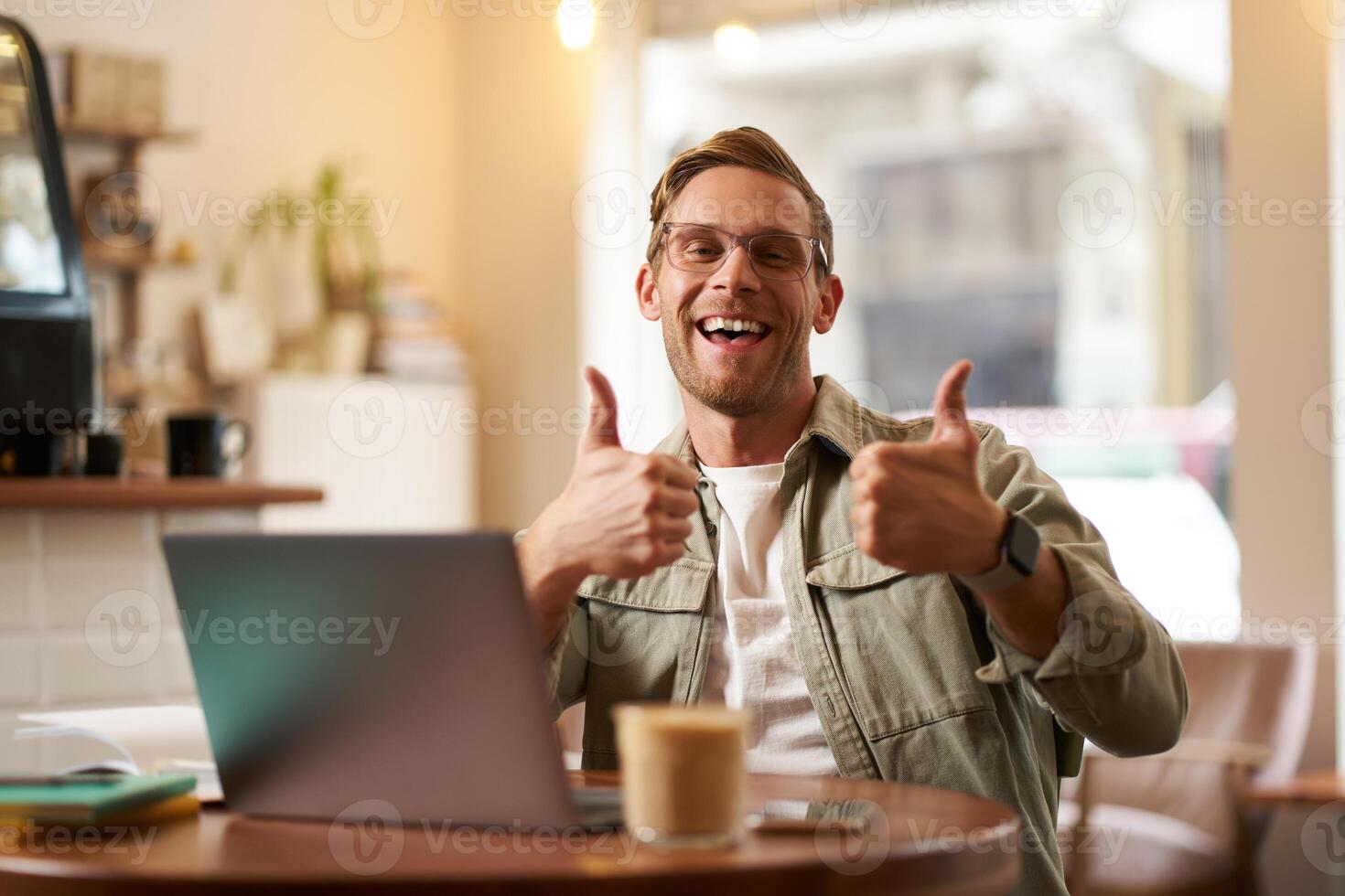 Portrait of handsome smiling man, shows thumbs up, sits in cafe with cup of coffee and laptop, approves smth good, recommends co-working space or online website photo