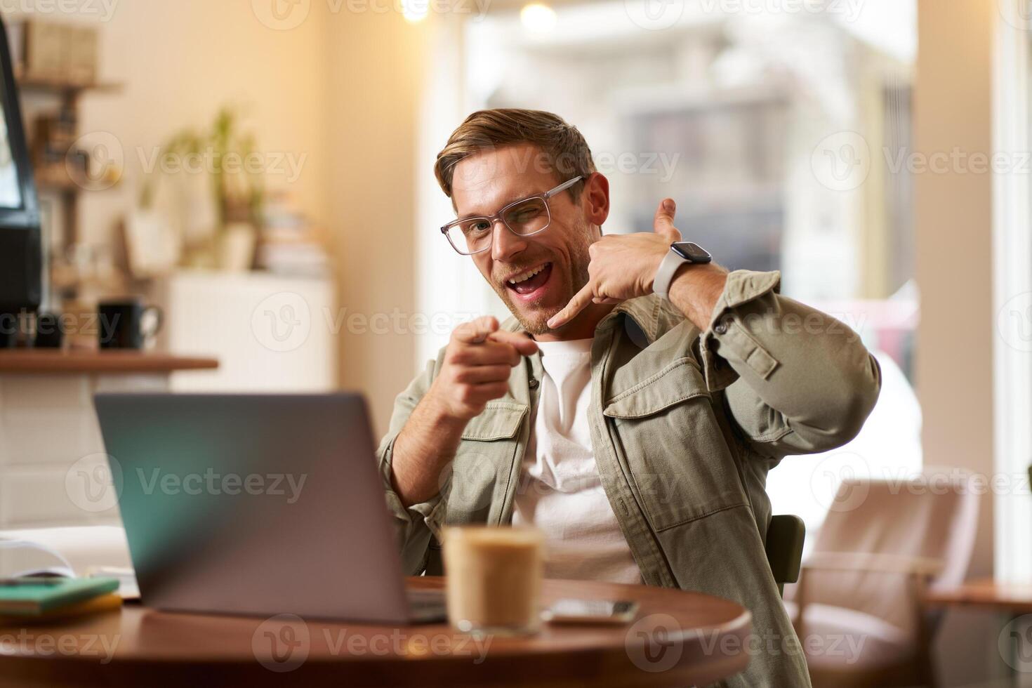Portrait of cheerful, flirty guy in glasses, sitting with laptop computer in cafe, showing phone call hand gesture and winking at you, giving his number photo