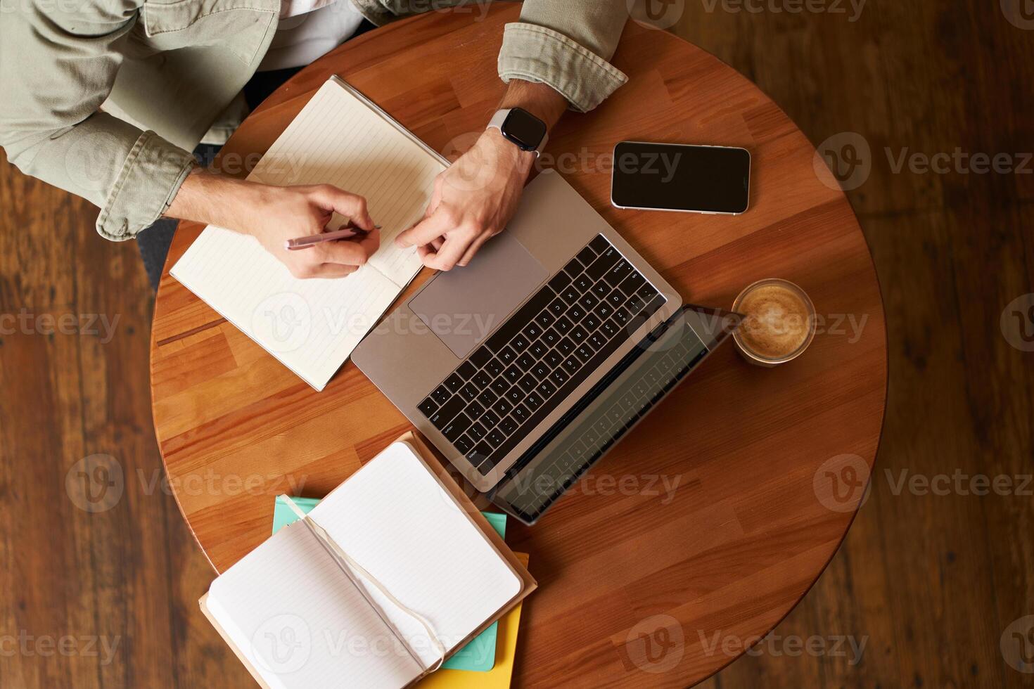Top aerial view of male hands taking notes, writing in notebook, man sitting at round desk in cafe, working on laptop, studying photo