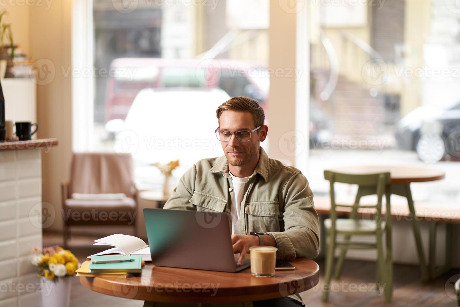 Image of young digital nomad, man in glasses sits in cafe, works from coffee shop, uses laptop in co-working space, wears glasses, drinks his beverage photo