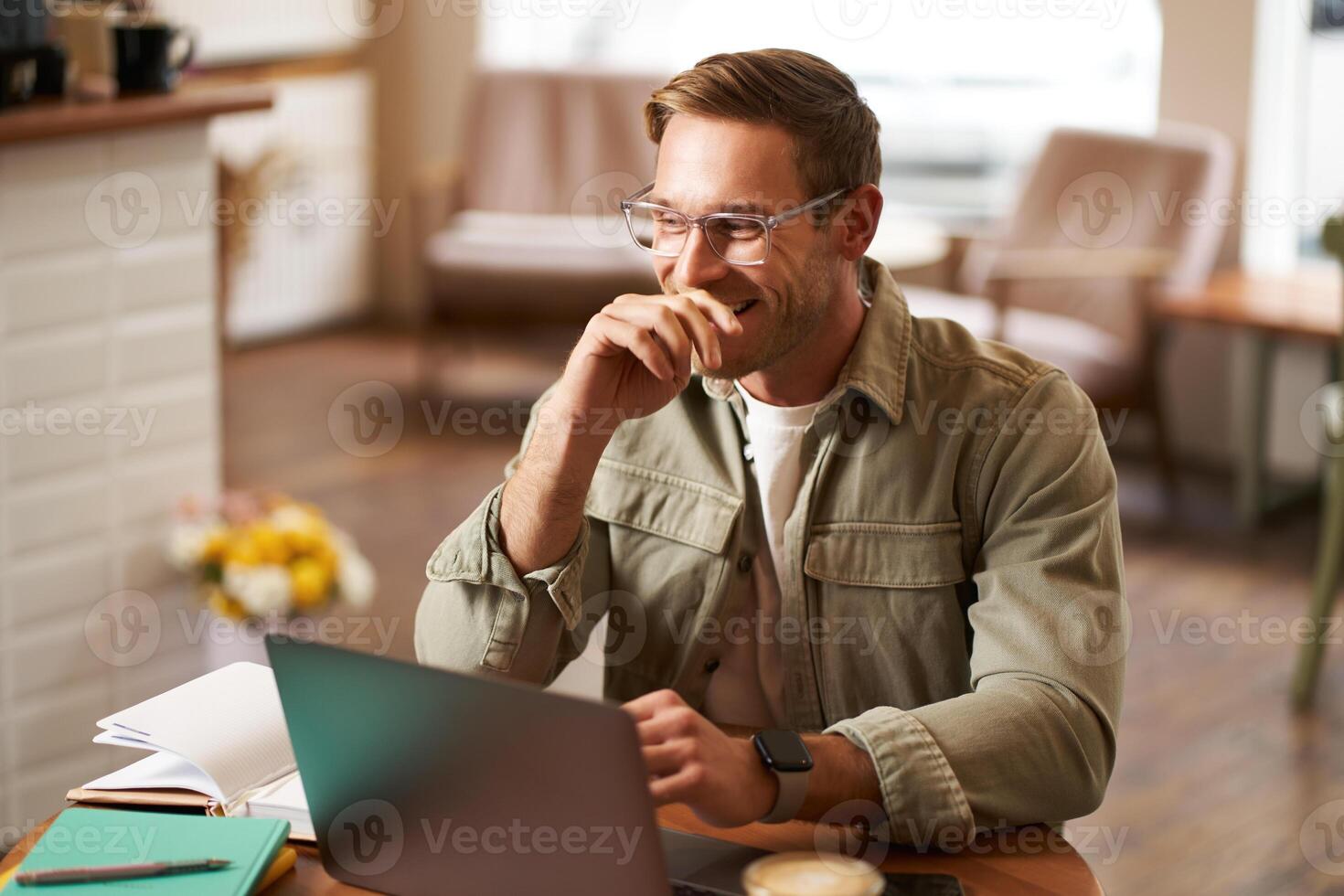 Image of young digital nomad, man in glasses sits in cafe, works from coffee shop, uses laptop in co-working space, wears glasses, drinks his beverage photo