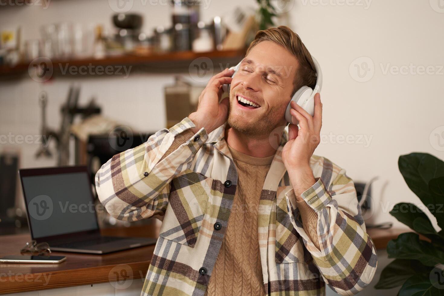 Handsome smiling young man, digital nomad, sitting in co-working open space, working in coffee shop, singing and listening to music in wireless headphones photo