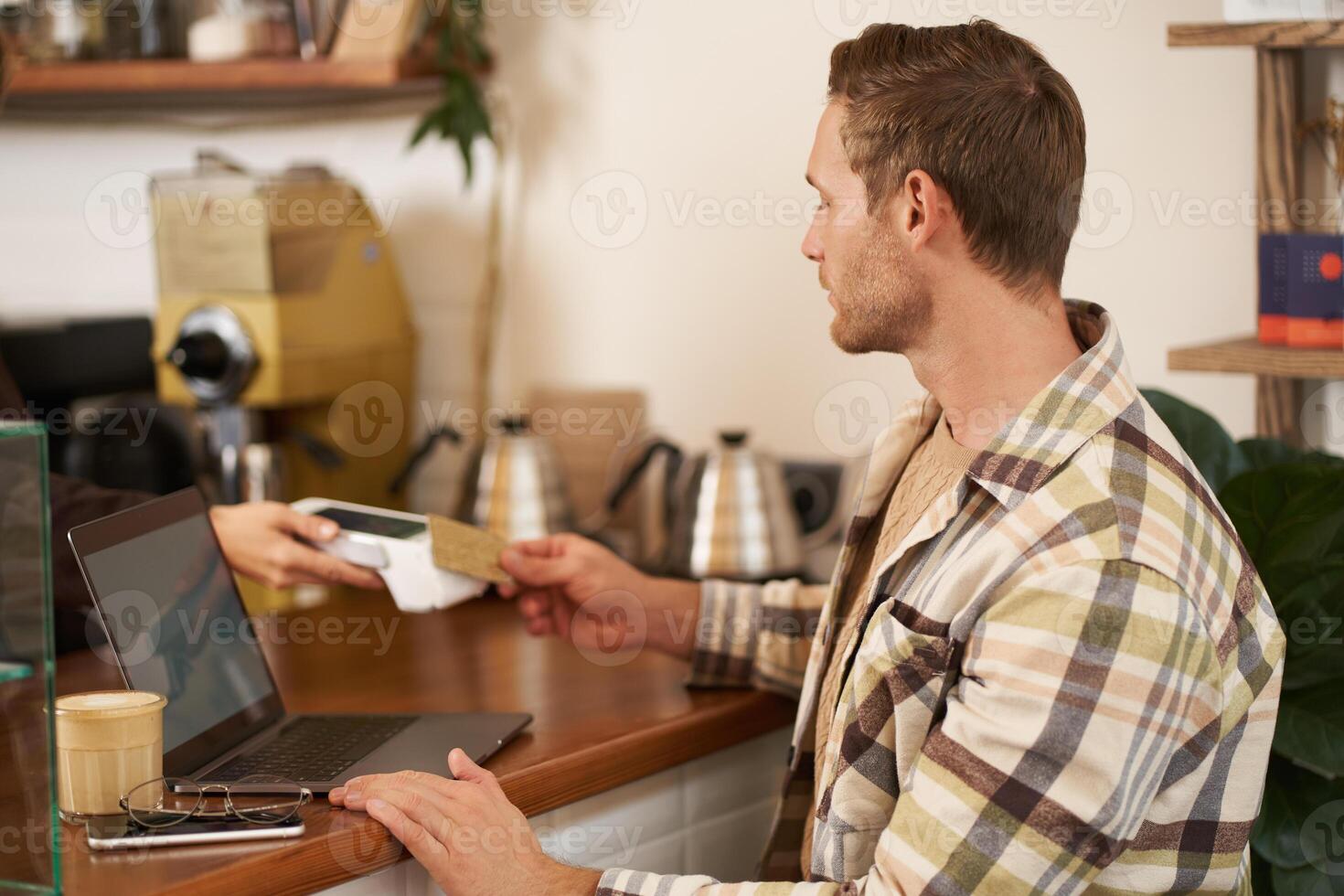 Portrait of freelancer, young man with laptop in coffee shop, paying for his order, pressing his credit card to the POS terminal, contactless payment concept photo