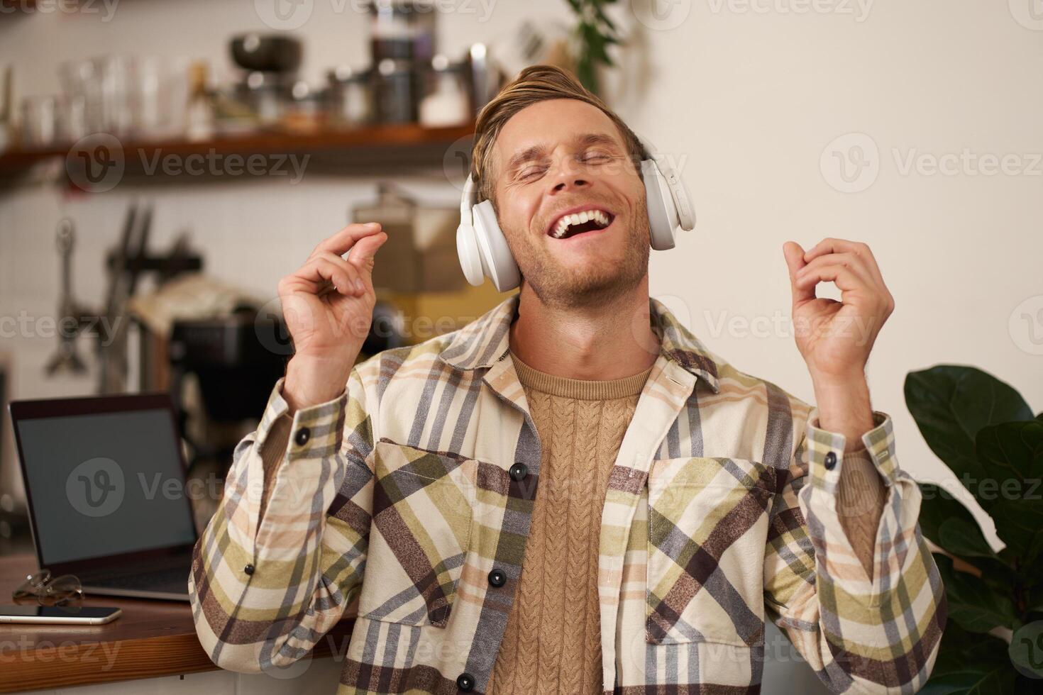 Handsome smiling young man, digital nomad, sitting in co-working open space, working in coffee shop, singing and listening to music in wireless headphones photo