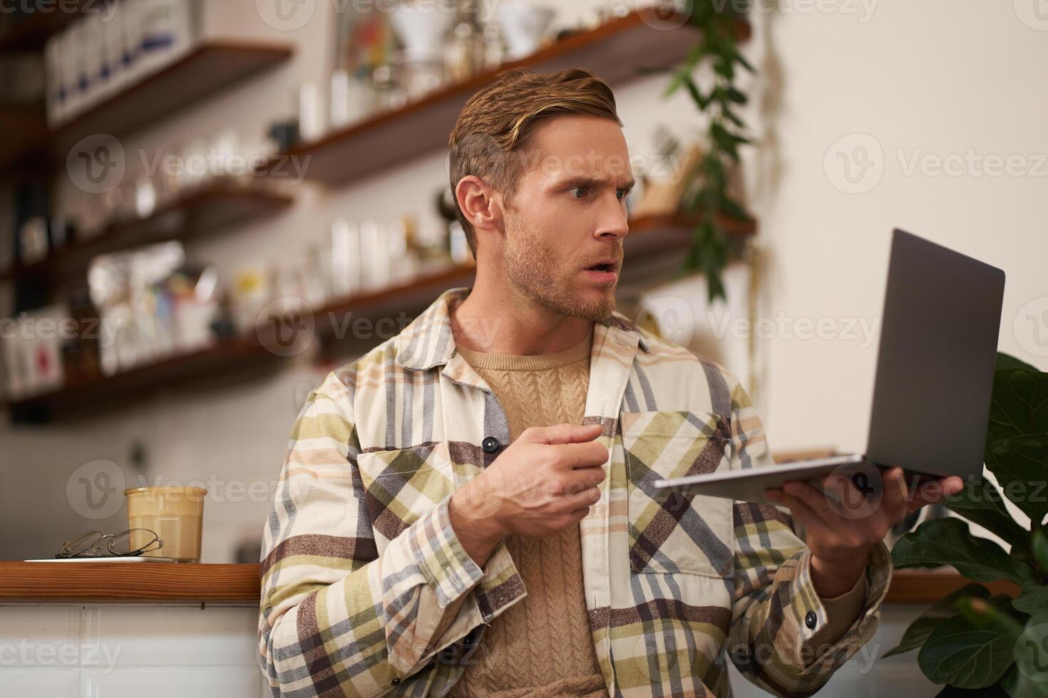 Portrait of man looking stressed and concerned at laptop screen, clench fist and frowns with angry face expression, sits in cafe with cup of coffee photo