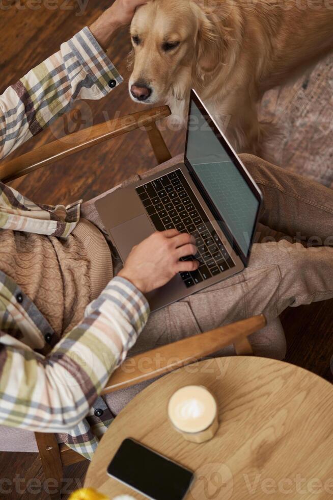 Vertical cropped photo of male hands and body, man petting the dog while working on laptop in cafe, drinking coffee