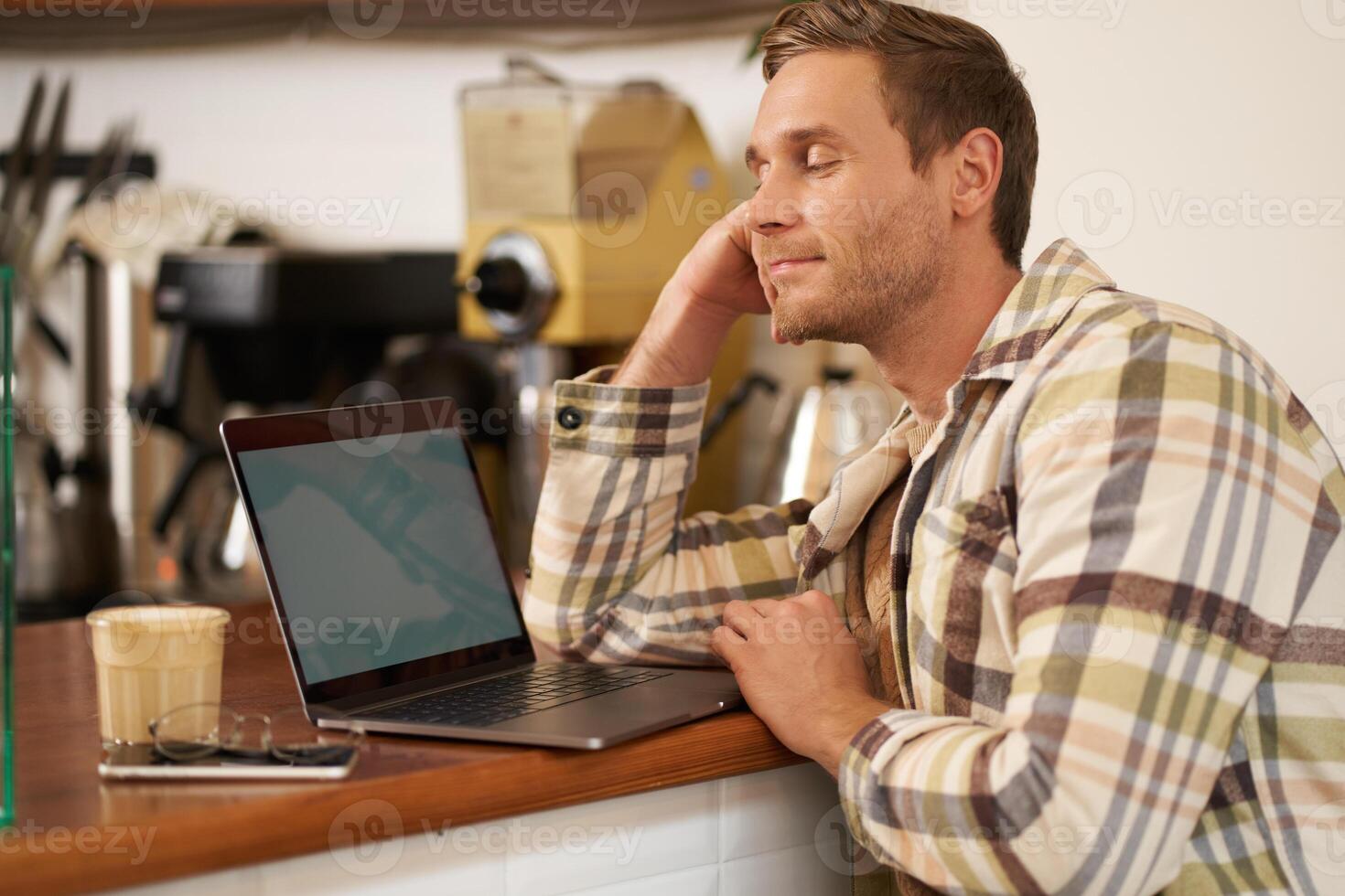 Close up portrait of handsome man, sitting at counter and looking at laptop screen with pleased smile, drinking coffee, browsing on the internet, watching something on computer photo