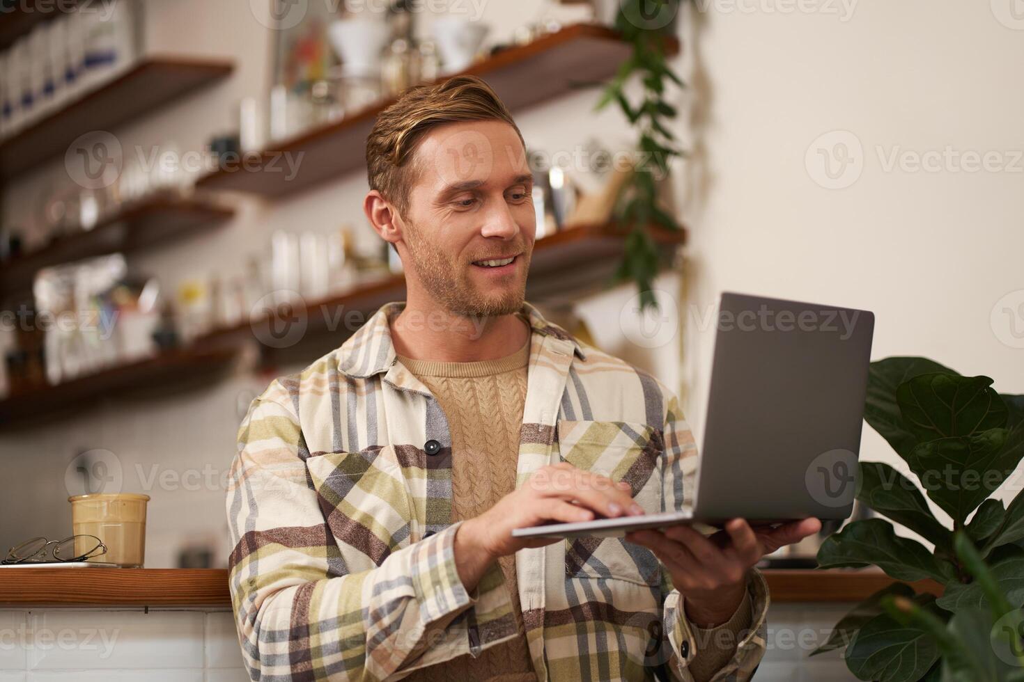 Portrait of young man working online, freelancer sitting in cafe, doing his project on laptop, drinking coffee, smiling while looking at screen photo