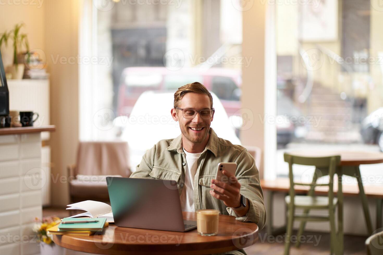 Portrait of handsome man in glasses, sits in cafe with laptop, smiles while reads a message on phone, checks his smartphone app, works from coffee shop photo