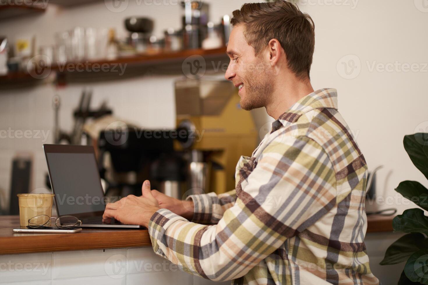 Portrait of handsome smiling man, sitting with laptop in a cafe, typing, working remotely from co-working space, drinking coffee, freelancing from public space photo