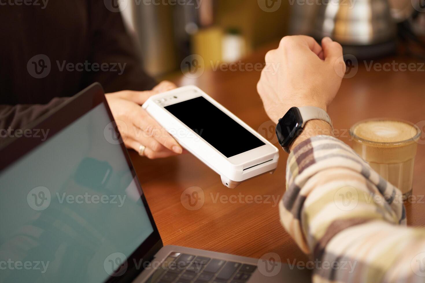 Close up shot of man paying with his watch, holding wrist near POS terminal for contactless payment, sitting in cafe co-working space with laptop photo