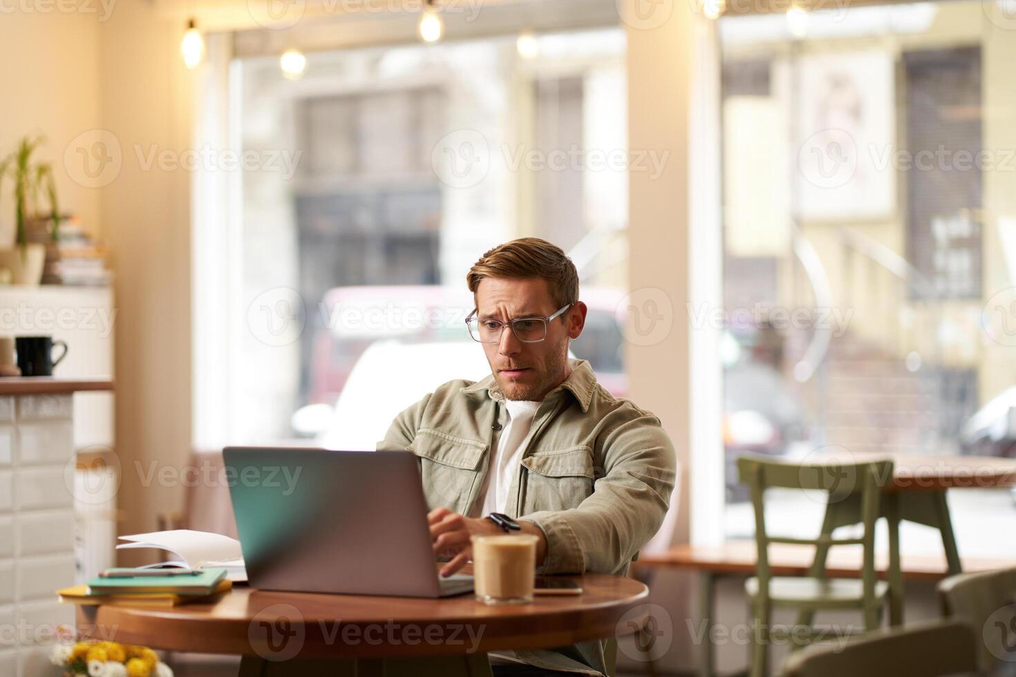 Image of young digital nomad, man in glasses sits in cafe, works from coffee shop, uses laptop in co-working space, wears glasses, drinks his beverage photo