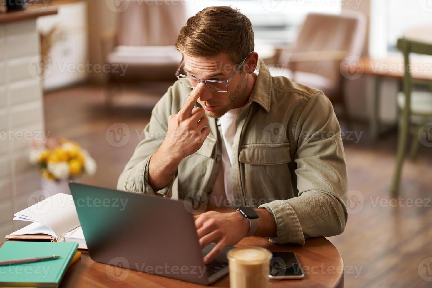 Image of young digital nomad, man in glasses sits in cafe, works from coffee shop, uses laptop in co-working space, wears glasses, drinks his beverage photo