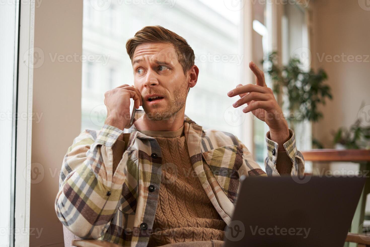 Portrait of serious businessman talking over the phone, sitting in chair in a cafe with laptop, working on project, discussing work-related issues on telephone, looks behind him photo