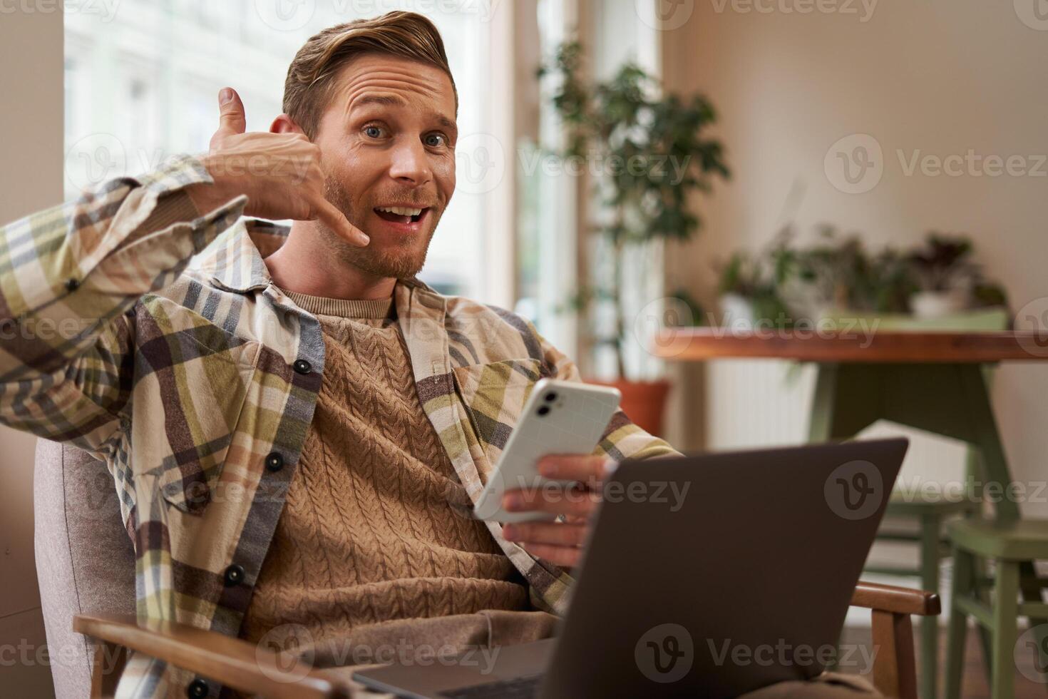 Portrait of funny handsome cafe visitor, young man with laptop and smartphone, showing mobile phone hand sign and smiling at camera, asking to give him a call, enquire over the telephone photo