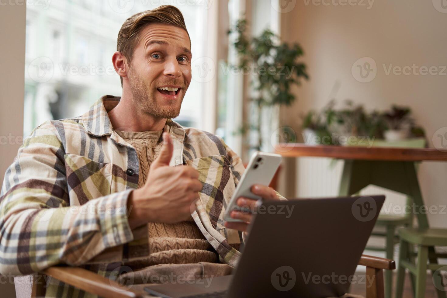 Happy cafe visitor, young man freelancer, working from coffee shop, sitting with phone and laptop, showing thumbs up and saying yes, approve smth good, gives positive feedback or review photo