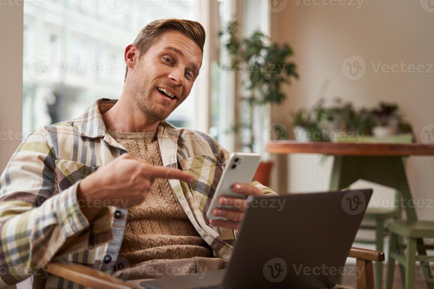 emocionado chico en cafetería, sentado con teléfono y señalando a pantalla con interés, recomendando un aplicación, utilizando móvil dispositivo y ordenador portátil en trabajo colaborativo espacio. concepto de Lanza libre y remoto espacio de trabajo foto