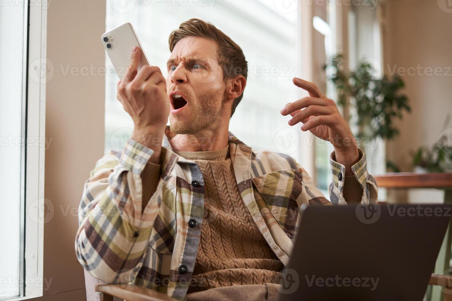 Portrait of an angry man shouting at his mobile phone. Frustrated cafe visitor arguing with someone on call, sitting with laptop near window, screaming at telephone photo