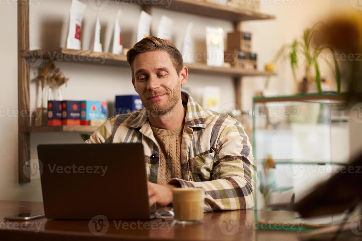 Portrait of handsome young digital nomad, man working in cafe on laptop, looking happy and pleased with his online project, sitting in co-working coffee shop photo