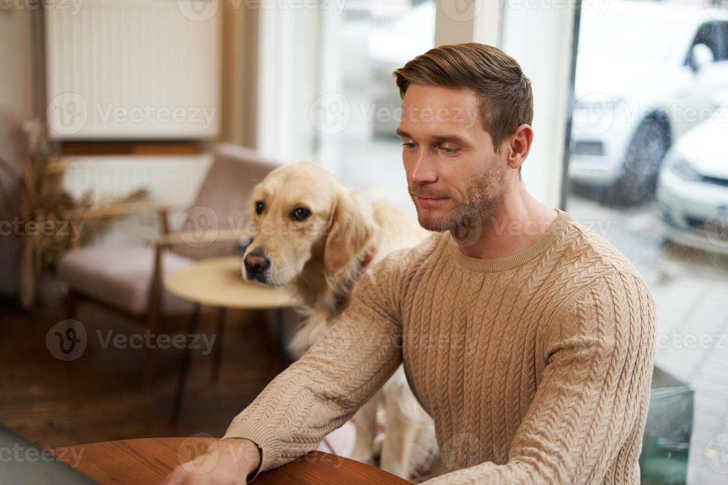 hermoso joven hombre, digital nómada sentado con su perro en un mascota amigable cafetería, trabajando en ordenador portátil mientras un dorado perdiguero relojes él foto