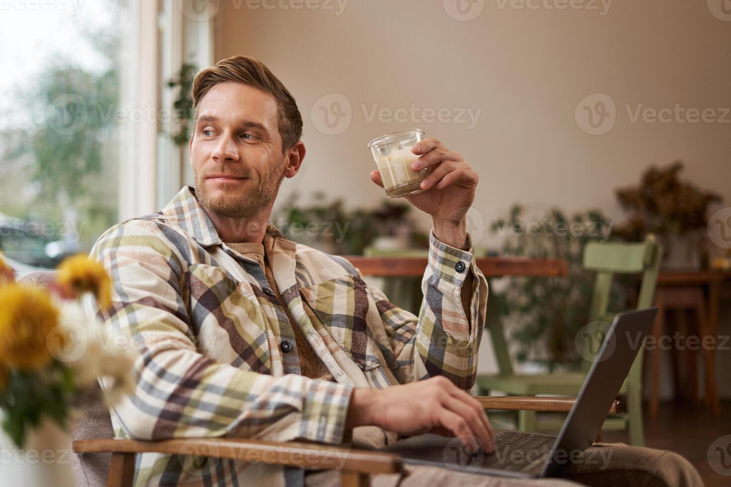 Portrait of handsome young man, working in cafe, sitting with laptop and coffee in hand, has remote job and using local co-working space instead of going to the office photo