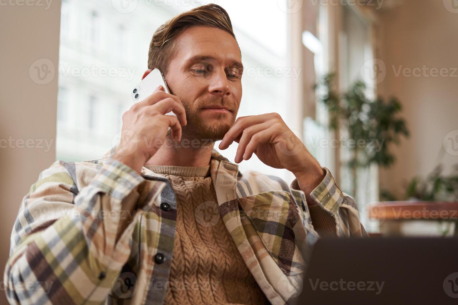 Image of young professional, cafe visitor working, sitting with laptop and talking on mobile phone to client. A guy making an enquiry buying something online, calling a friend photo