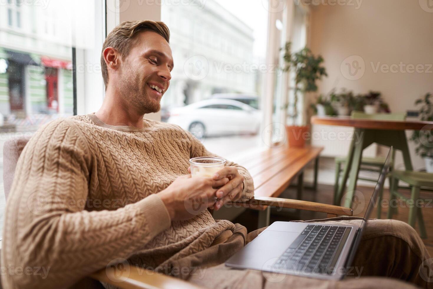 Portrait of handsome young man sits in cafe, drinks coffee and watches on laptop, looking at screen with happy smile, relaxing in co-working space photo