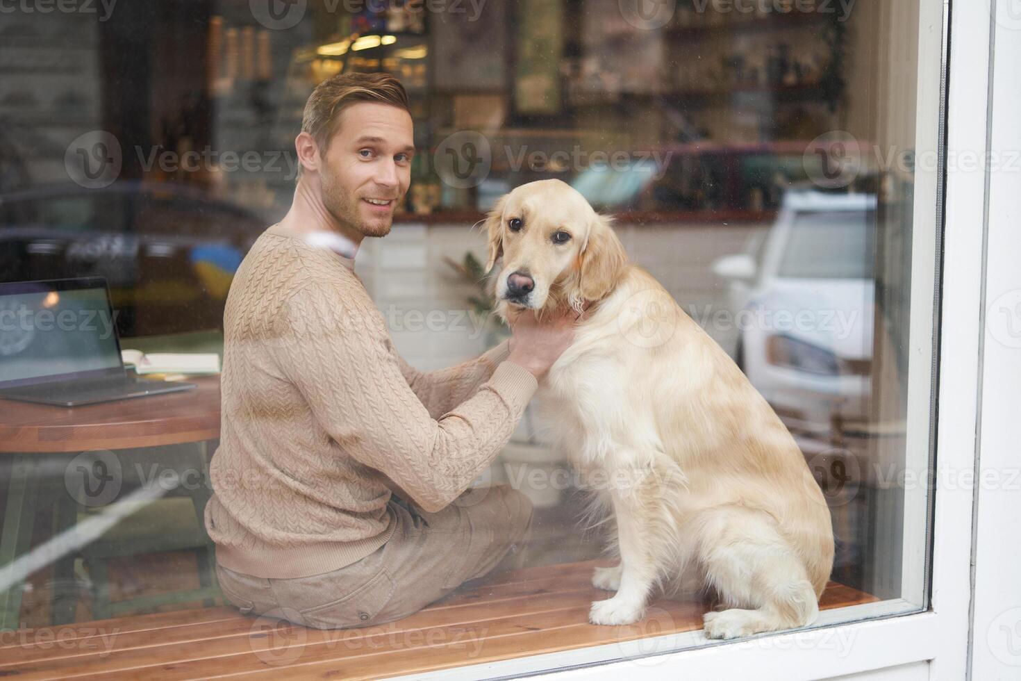 Outdoor shot of a pet-friendly cafe where man sits near the window with his fluffy golden retriever and smiles at camera. Coffee shop visitor with a dog photo