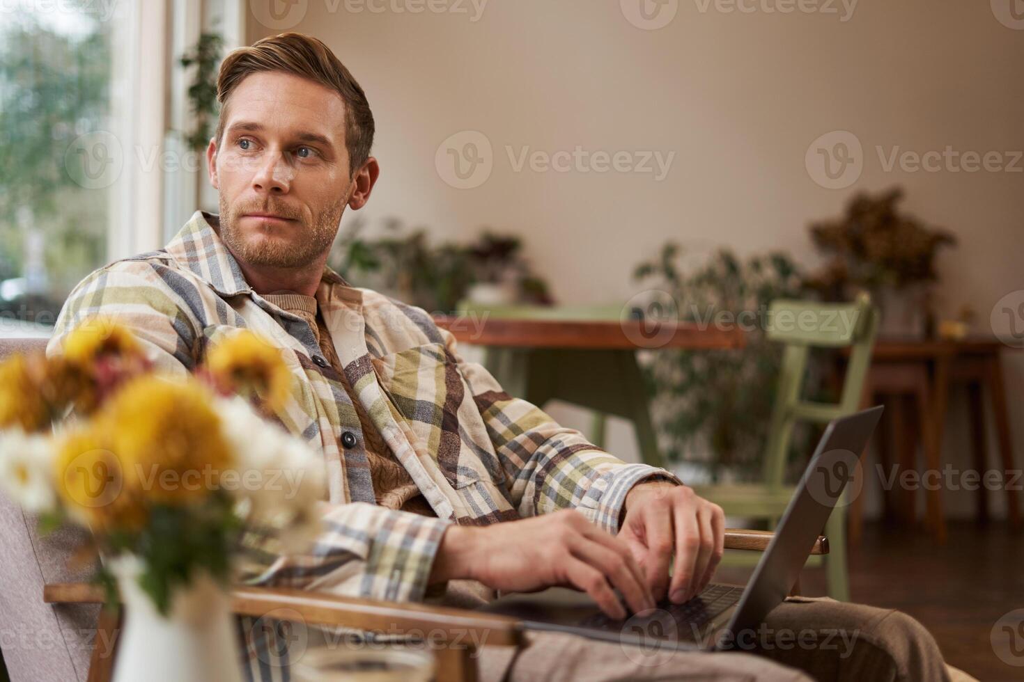 Portrait of handsome young man, working in cafe, sitting with laptop and coffee in hand, has remote job and using local co-working space instead of going to the office photo